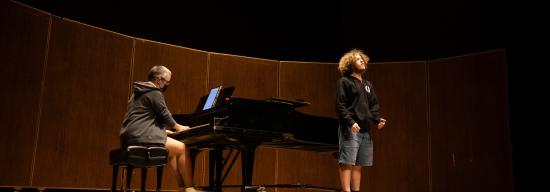 Male student singing on stage with a pianist behind him playing music