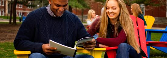 Two students reading a book while sitting on chairs outside